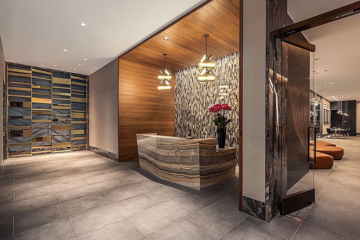 This sophisticated lobby or reception area features a striking stone-clad desk as its centerpiece, set against a textured mosaic wall and warm wood-paneled ceiling. The space is accented by modern geometric pendant lighting and a decorative gold-and-blue wall installation, creating a luxurious and welcoming atmosphere. The perspective is a wide-angle shot from the entrance, showcasing the high-end finishes and seamless transition into the adjacent lounge area.