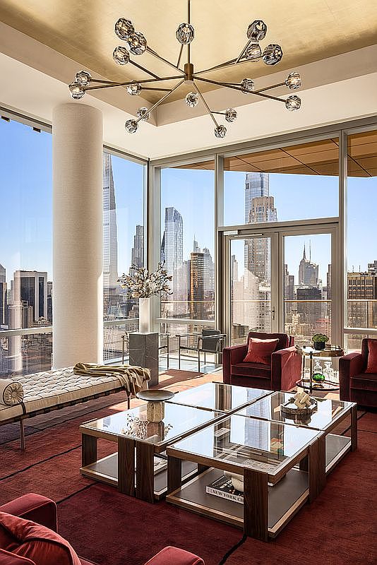 This sophisticated high-rise living room features floor-to-ceiling windows that offer a breathtaking panoramic view of the New York City skyline. The space is anchored by a rich, deep red area rug, a modern modular glass coffee table, and plush velvet seating, all illuminated by a striking, multi-armed contemporary chandelier. The combination of warm metallic accents, sleek architectural lines, and the dramatic urban backdrop creates an atmosphere of refined luxury and exclusivity.
