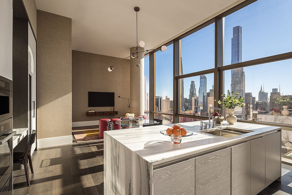 This sophisticated, high-end kitchen features a stunning waterfall marble island and sleek, wood-textured cabinetry, seamlessly blending into an open-concept living area. Floor-to-ceiling windows provide a breathtaking, panoramic view of the New York City skyline, bathing the space in natural light. The modern aesthetic is complemented by a minimalist pendant light fixture and dark hardwood flooring, creating an atmosphere of urban luxury.