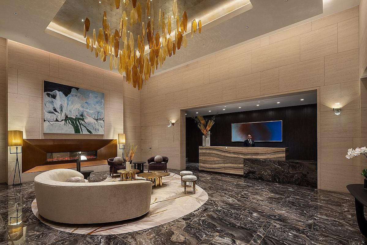 This luxurious lobby features a grand, high-ceilinged space with a striking gold-leaf chandelier and polished dark marble flooring. A curved, cream-colored sofa sits atop a circular rug in front of a modern fireplace, while a sleek reception desk with a marble facade anchors the background. The overall aesthetic is sophisticated and high-end, conveying a sense of exclusivity and professional hospitality.