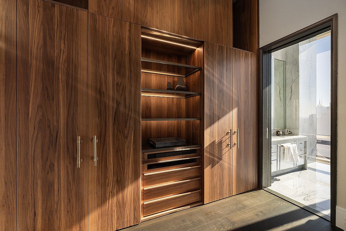 This sophisticated walk-in closet features rich, dark walnut cabinetry with integrated LED lighting that highlights the open shelving and organized drawer units. The seamless design includes sleek metal hardware and leads directly into a bright, marble-clad bathroom visible through a doorway. The overall aesthetic is one of high-end luxury, characterized by clean lines, premium materials, and a warm, inviting atmosphere.
