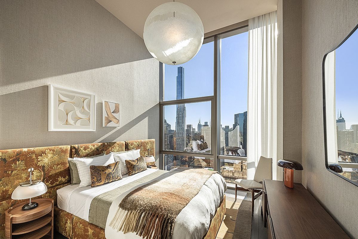 This sophisticated primary bedroom features a plush, floral-patterned headboard and a cozy, layered bed set against a backdrop of floor-to-ceiling windows that offer a breathtaking, high-altitude view of the New York City skyline. The room is accented by a modern spherical pendant light, a sleek wooden nightstand, and a minimalist dresser, creating a luxurious and serene urban retreat. The cinematic perspective captures the contrast between the warm, textured interior and the cool, expansive cityscape outside.