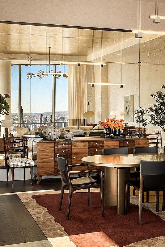 This sophisticated dining room features a warm, mid-century modern aesthetic with a rich wooden sideboard and a sleek, circular table surrounded by dark leather chairs. A striking, multi-armed chandelier hangs above, while a large mirrored wall reflects the expansive city skyline view visible through the floor-to-ceiling windows. The space is grounded by a deep, textured rug, creating an atmosphere of refined luxury and urban elegance.