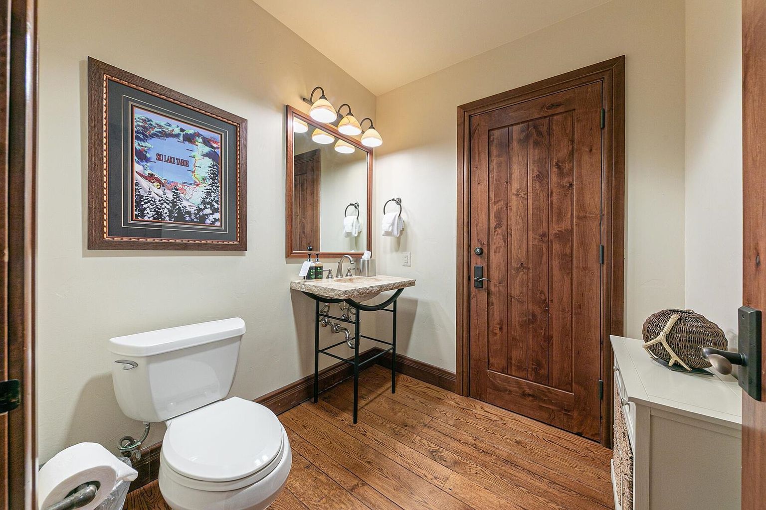 This is a cozy guest bathroom featuring a wooden door and hardwood floors, creating a warm and inviting atmosphere. The bathroom includes a toilet, a unique stone sink with a metal stand, and a framed picture on the wall, adding a touch of personality. A decorative basket sits atop a white cabinet, enhancing the room's charm.