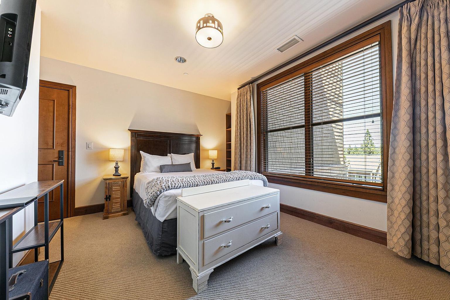 This is a cozy guest bedroom featuring a dark wood headboard and a white dresser at the foot of the bed. The room is well-lit with a ceiling light and bedside lamps, and a large window with blinds provides natural light. The neutral color palette and carpet create a warm and inviting atmosphere.