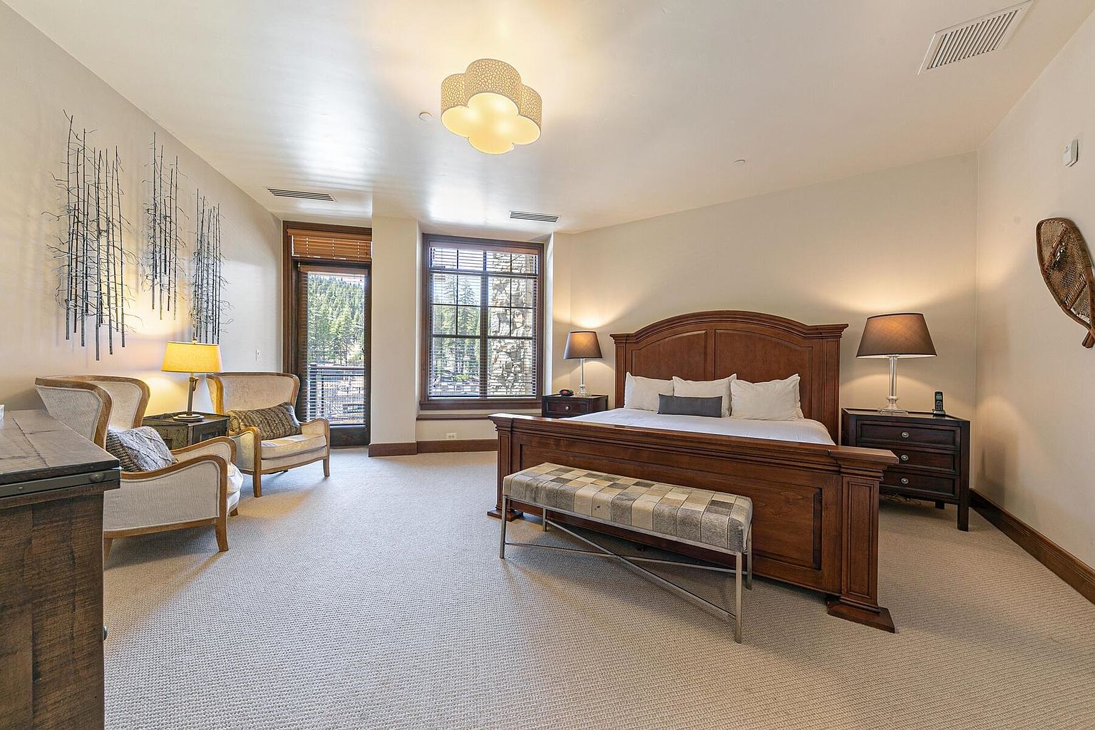 This is a spacious primary bedroom featuring a large wooden bed with white linens and a plaid bench at the foot. The room is well-lit with natural light from the windows and complemented by bedside lamps. The decor includes artwork and comfortable seating, creating a warm and inviting atmosphere.