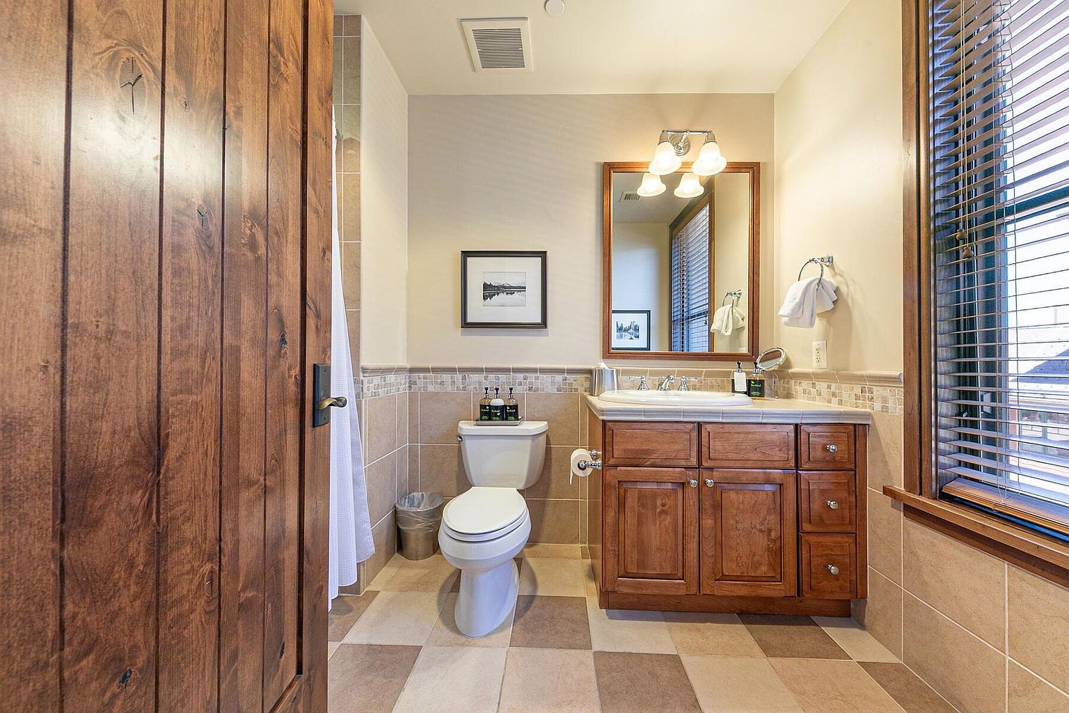 This is a well-lit bathroom featuring a wooden vanity with a white countertop and sink, complemented by a framed mirror and decorative lighting. A toilet sits adjacent to a tiled wall, and the floor is covered in a checkered pattern of neutral-toned tiles. The overall impression is clean and functional, with a touch of rustic charm.