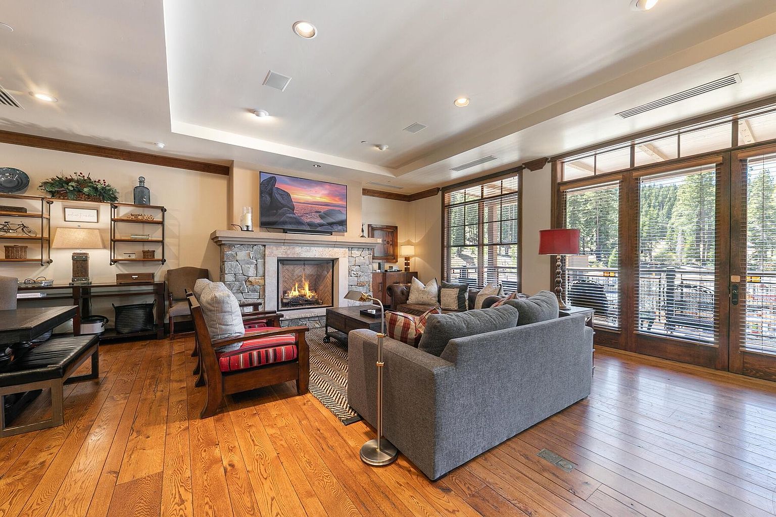 This is an interior shot of a living room featuring hardwood floors, a stone fireplace with a mounted television, and comfortable seating including a gray sofa and wooden chairs with striped cushions. Large windows with blinds provide natural light and a view of the outdoors. The room exudes a cozy and inviting atmosphere, perfect for relaxation and entertainment.