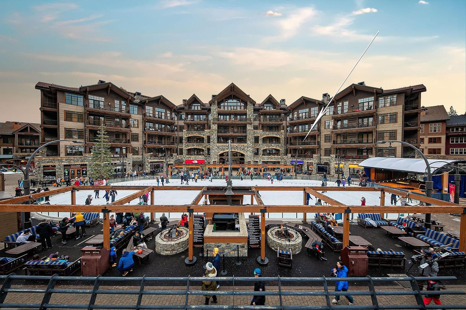 This image showcases a vibrant outdoor community space featuring an ice skating rink surrounded by seating areas with fire pits. The area is framed by a large, multi-story building with a rustic architectural style, suggesting a resort or residential complex. The scene conveys a sense of community and leisure, highlighting the amenities available to residents or visitors.