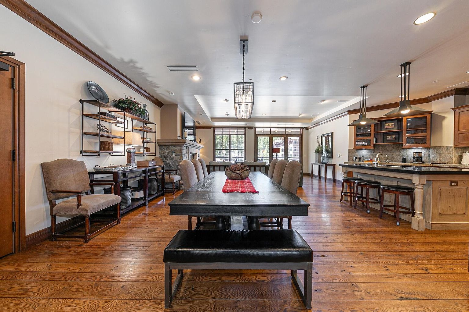 This is an interior shot of a dining room featuring a long, dark wood table with a bench and chairs. A decorative centerpiece and red runner adorn the table. The room also includes a shelving unit with decorative items, a fireplace, and a partial view of the kitchen with bar stools, creating a warm and inviting atmosphere.