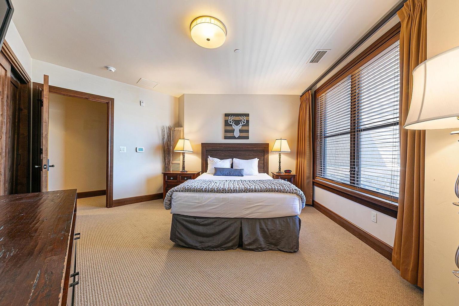 This is a cozy primary bedroom featuring a king-size bed with a wooden headboard, flanked by two nightstands with lamps. A large window with blinds and curtains provides natural light. The room is carpeted and has a warm, inviting feel, perfect for relaxation.