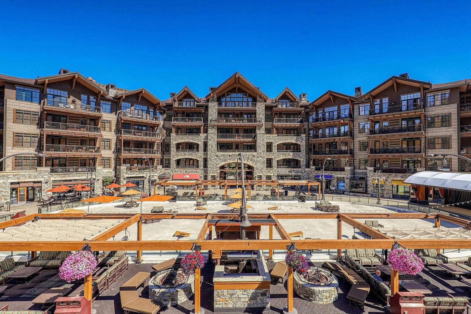 This aerial view showcases a luxurious multi-story building complex with stone and wood facade details, designed in a mountain resort style. The focal point is a central outdoor gathering area featuring fire pits, plush seating, and wooden pergola structures, suggesting a high-end amenity space. The clear blue sky enhances the image, giving a sense of place and spaciousness.