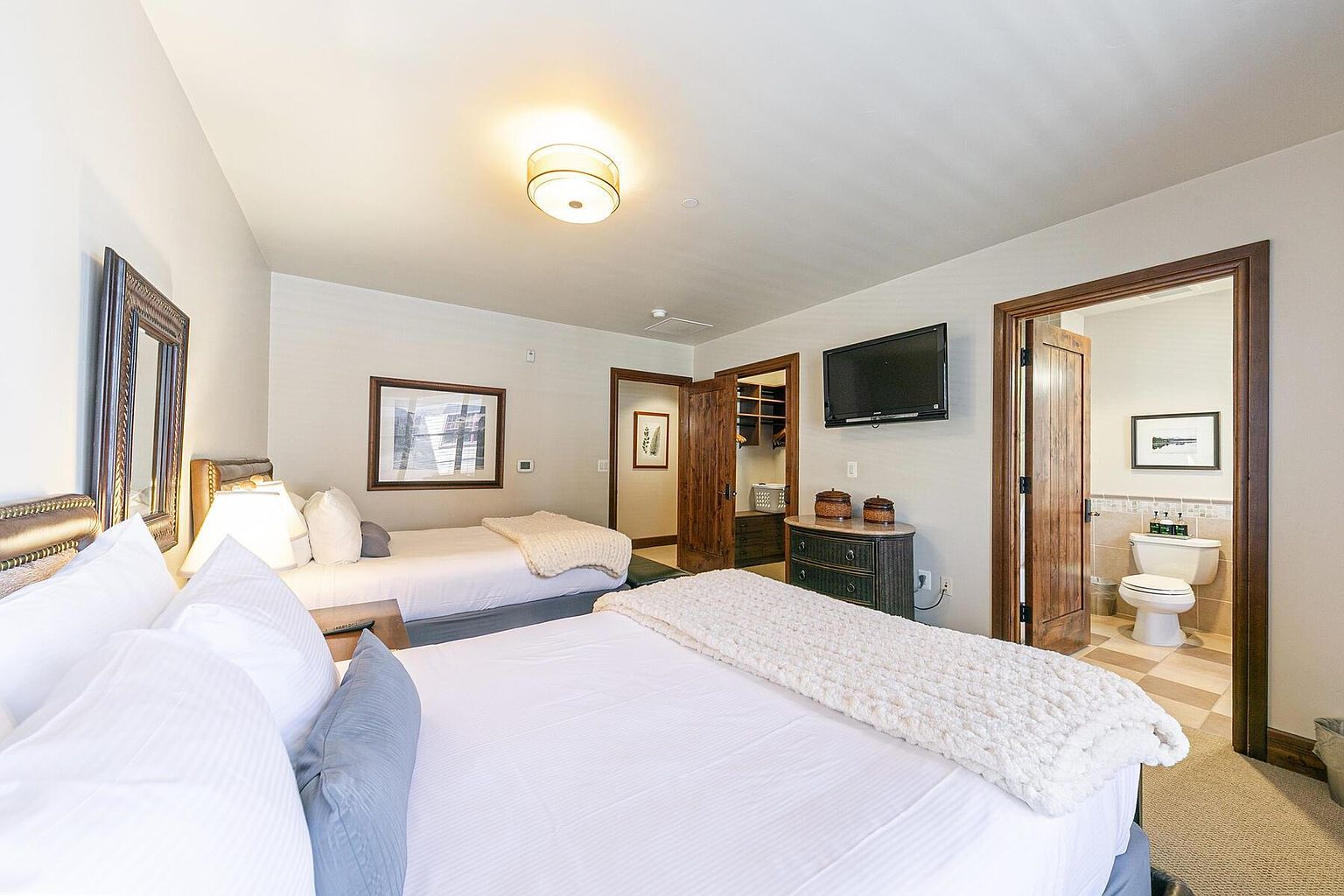 This is a well-lit guest bedroom featuring two twin beds with white linens and decorative pillows. The room includes a mounted television, a dresser with decorative items, and doorways leading to a closet and a bathroom. The overall impression is clean, comfortable, and inviting, suitable for guests or family members.
