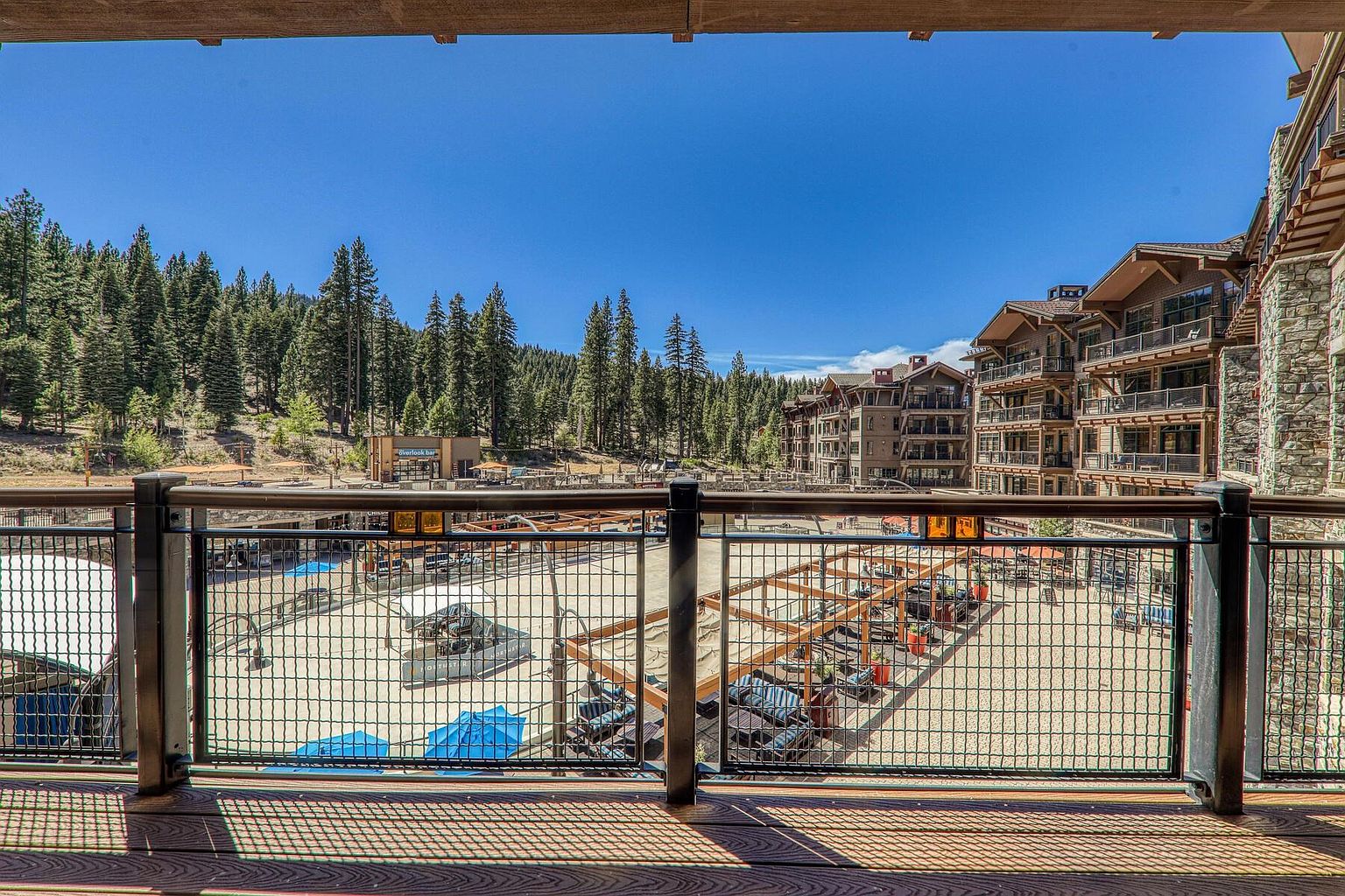 This image showcases a spacious balcony overlooking a resort-style outdoor area. The balcony features a wooden deck and a metal railing, providing a clear view of the pool, lounge chairs, and surrounding buildings. The scene is bathed in sunlight, creating a warm and inviting atmosphere, perfect for relaxation and entertainment.