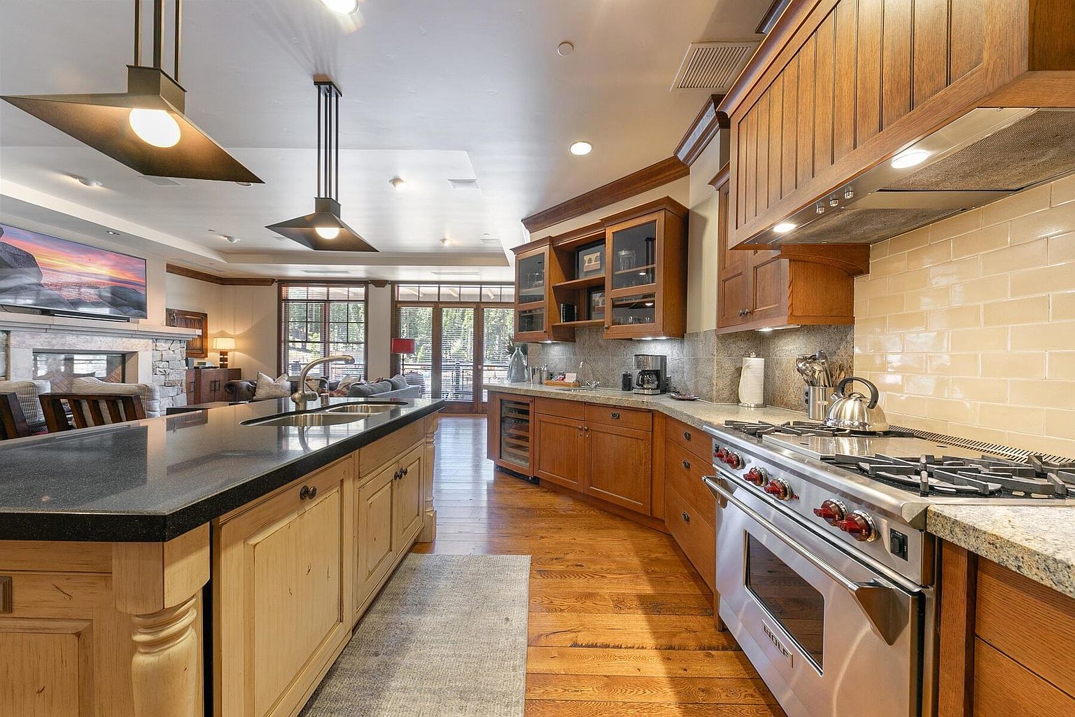 This is a spacious kitchen featuring wooden cabinetry, granite countertops, and stainless steel appliances. A large island with a dark countertop provides ample workspace and seating. The kitchen seamlessly transitions into a living area with a fireplace and large windows, creating an open and inviting atmosphere.