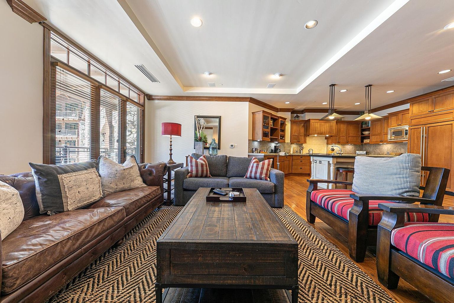 This is an interior shot of a living room that flows into a kitchen area. The living room features a leather sofa, a gray sofa, and two armchairs, all arranged around a dark wood coffee table on a patterned rug. Large windows provide natural light, and the kitchen in the background has wooden cabinetry and modern lighting fixtures.