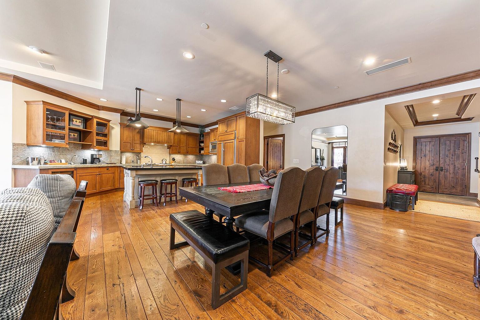 This interior shot showcases a dining area adjacent to a kitchen, featuring a large wooden dining table with upholstered chairs and a bench. A decorative chandelier hangs above the table, and the hardwood floors add warmth to the space. The open layout and natural light create an inviting atmosphere.