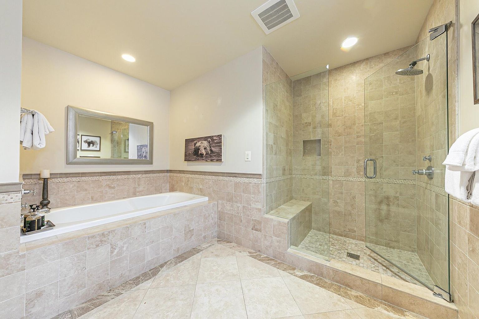 This is a well-lit primary bathroom featuring a large soaking tub with tile surround and a separate glass-enclosed shower. The bathroom has neutral-toned tile flooring and walls, creating a spa-like atmosphere. A framed mirror hangs above the tub, and artwork adorns the walls, adding a touch of personality.