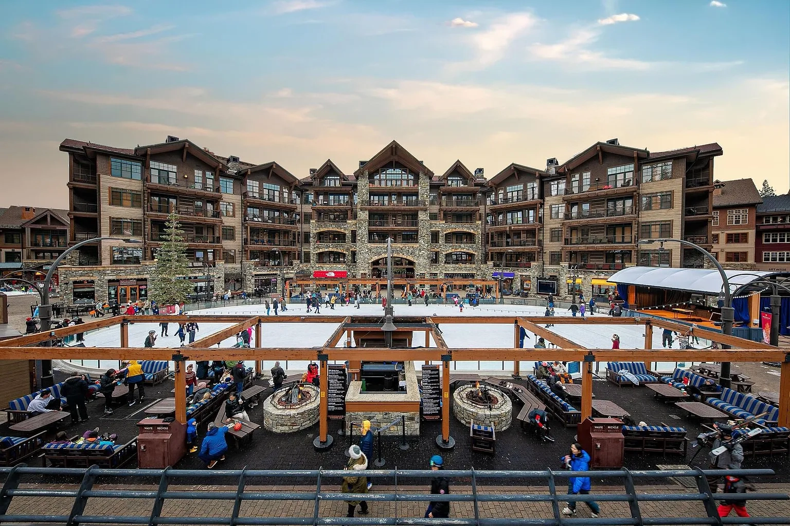 This image showcases a large, multi-story building that would be ideal for a ski resort or vacation property. The building has a rustic yet luxurious feel, with stone and wood accents throughout the exterior. In front of the building, there is an ice skating rink surrounded by seating areas.