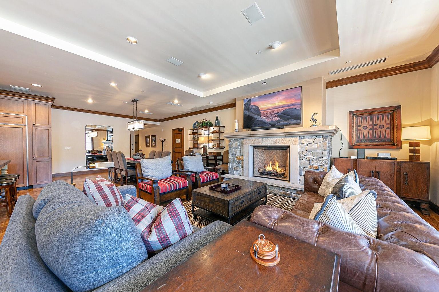 This is an interior shot of a living room featuring a stone fireplace with a television mounted above it. The room is furnished with a large leather sofa, a gray sofa with plaid pillows, and wooden armchairs. A wooden coffee table sits in the center of the room, and a dining area is visible in the background, creating an open-concept living space.
