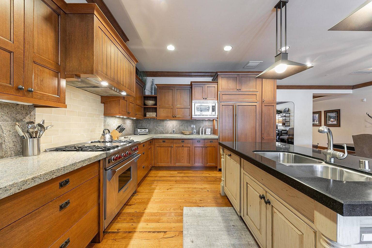 This is a well-appointed kitchen featuring wooden cabinetry, granite countertops, and stainless steel appliances. A large island with a dark countertop provides ample workspace and a sink. The hardwood floors and warm lighting create an inviting and functional space.
