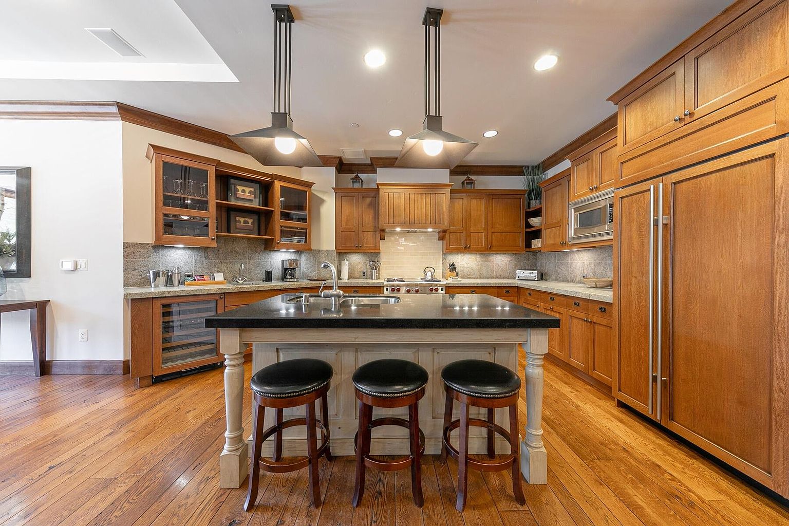 This is a warm and inviting kitchen featuring wood cabinetry, granite countertops, and stainless steel appliances. A central island with bar seating is illuminated by pendant lights, and the hardwood floors add to the room's cozy atmosphere. The kitchen is well-lit and appears to be spacious and functional.