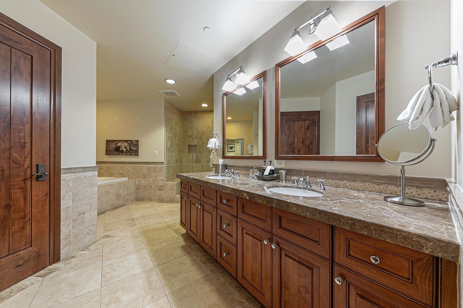 This is a well-appointed primary bathroom featuring a double vanity with a brown marble countertop and dark wood cabinetry. Two large mirrors are mounted above the sinks, illuminated by modern light fixtures. The bathroom includes a tiled floor and a separate tub and shower area, creating a luxurious and functional space.