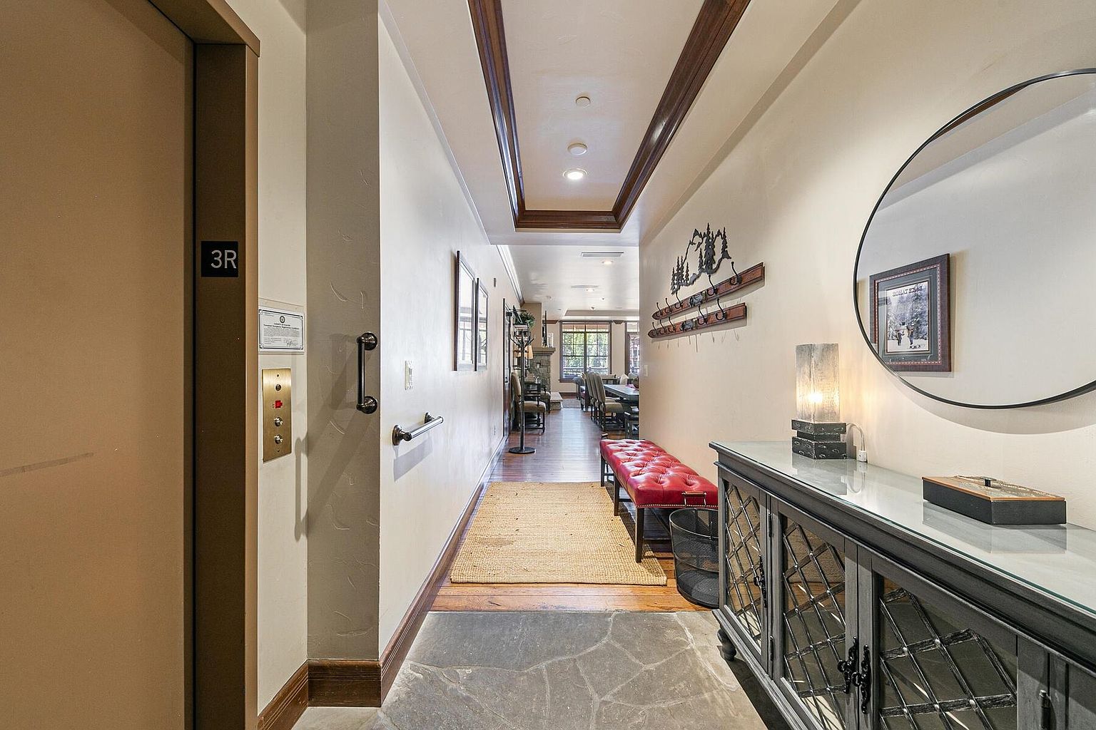 The image showcases a well-lit hallway with a blend of modern and rustic design elements. An elevator is visible on the left, while a red leather bench and a decorative console table with a lamp and mirror adorn the right side of the hallway. The flooring transitions from stone to wood, leading towards a dining area in the distance, creating a welcoming and stylish entrance to the living space.
