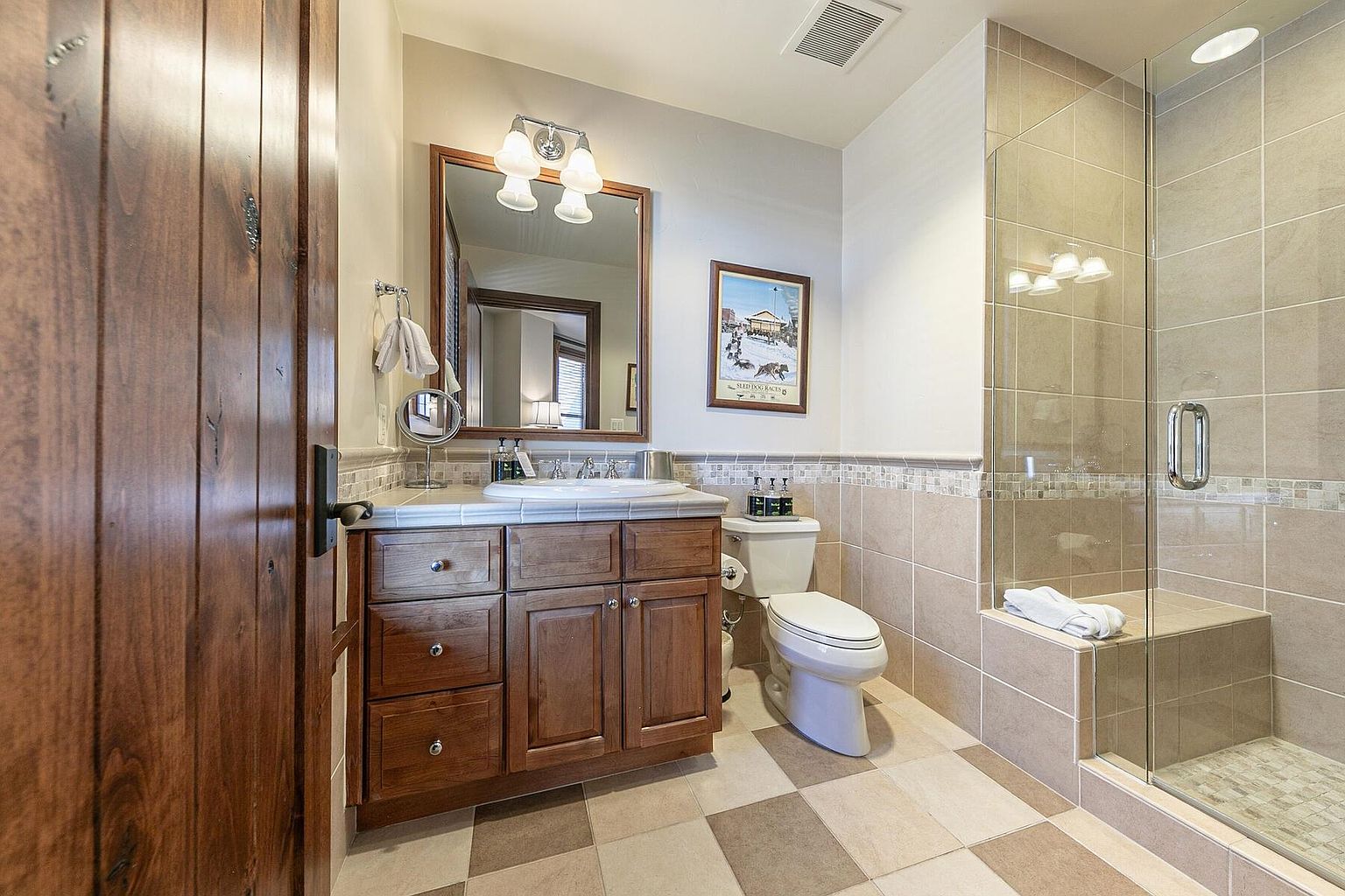 This is a well-maintained bathroom featuring a wooden vanity with a white countertop and sink, complemented by a framed mirror and overhead lighting. A toilet is positioned next to the vanity, and a glass-enclosed shower with tiled walls and a built-in bench is visible. The floor is tiled in a checkered pattern, adding a touch of visual interest to the space.
