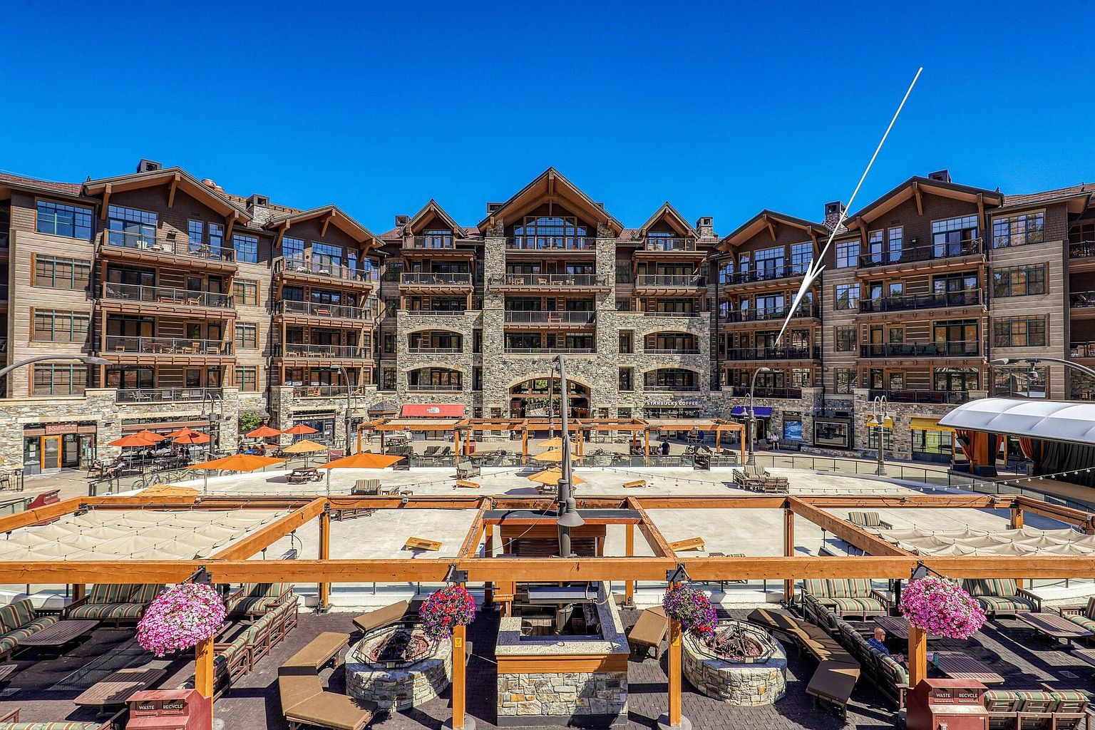 This image showcases a luxurious outdoor patio area, likely part of a resort or high-end condominium complex. The space features multiple seating areas with comfortable lounge furniture, fire pits, and overhead wooden structures adorned with hanging flower baskets. The backdrop includes a multi-story building with balconies and stone accents, creating an inviting and upscale atmosphere.