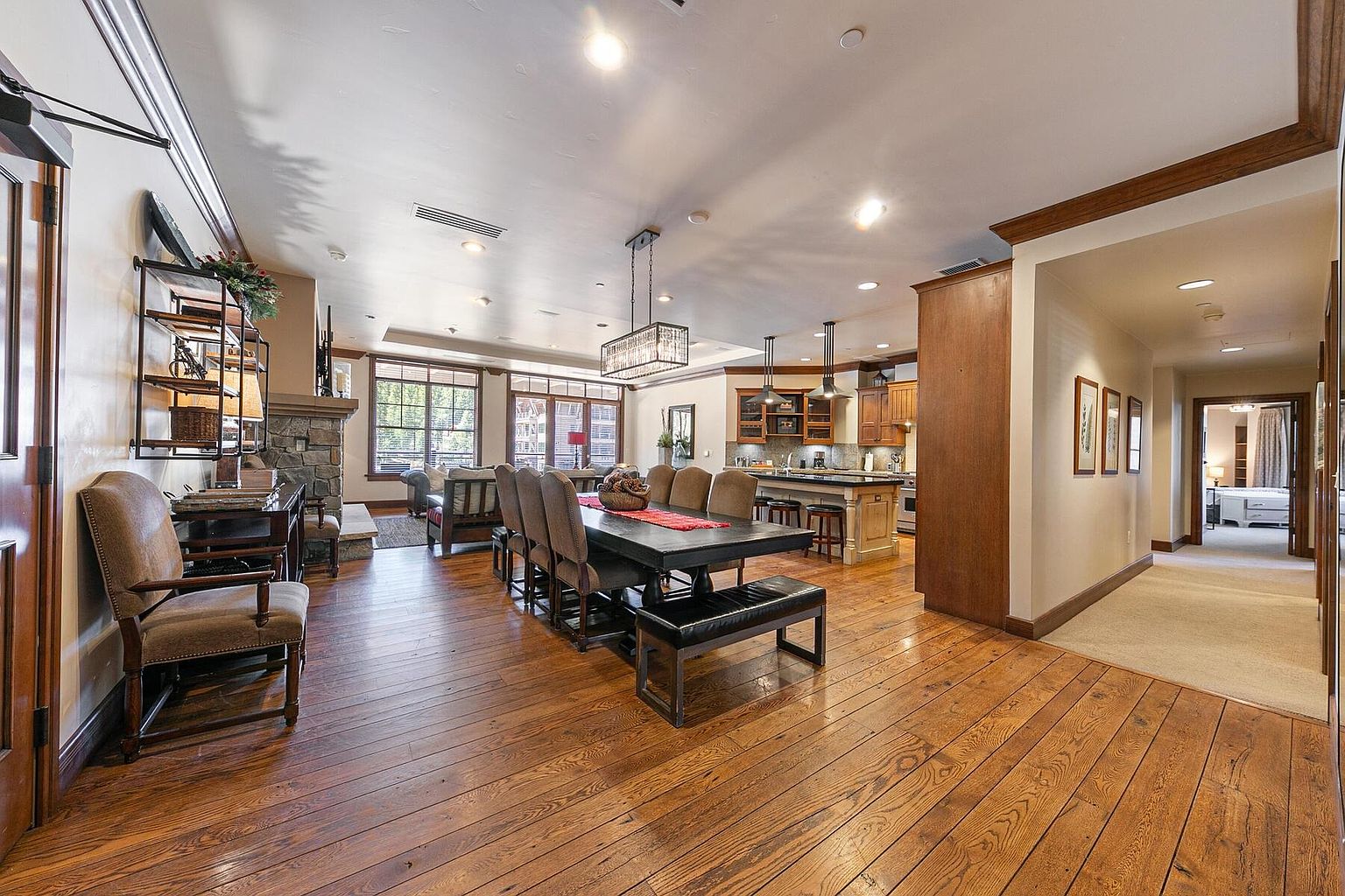 This expansive living area showcases a seamless blend of living, dining, and kitchen spaces, all unified by rich hardwood flooring. The dining area features a large table with bench seating, while the living room offers comfortable seating near a stone fireplace. The open-concept design enhances the sense of spaciousness and connectivity, ideal for entertaining and family living.