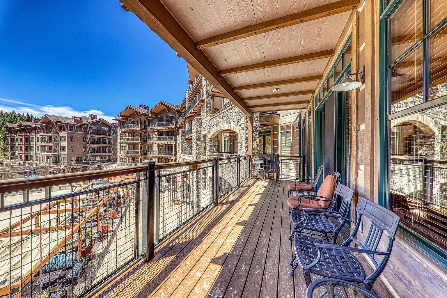 This image showcases a spacious balcony with wooden flooring and metal railings, offering a view of the surrounding buildings and outdoor area. The balcony is furnished with comfortable chairs and a small table, creating an inviting space for relaxation. The architecture features a blend of stone and wood, adding to the property's charm and appeal.