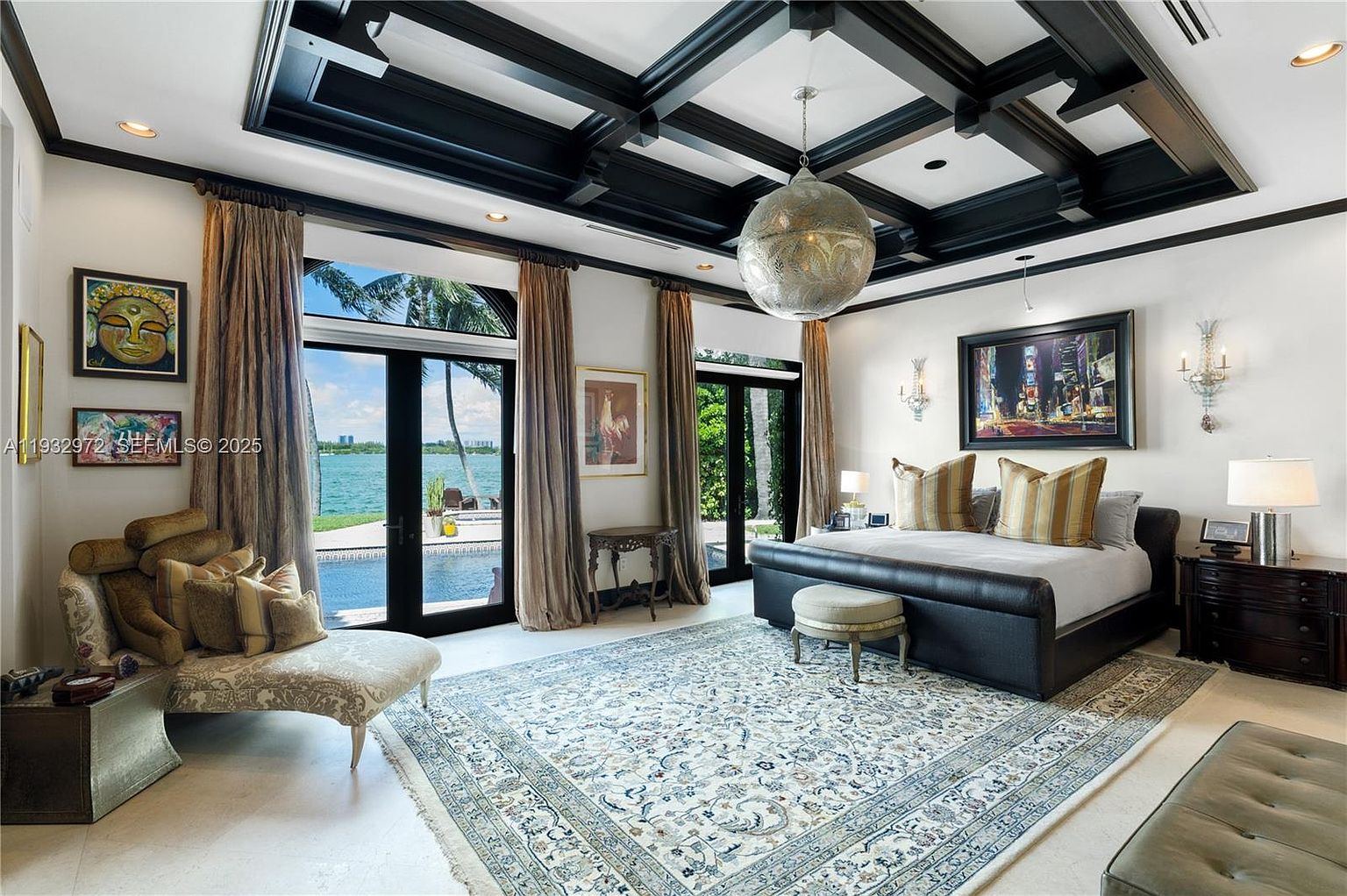 This luxurious primary bedroom features a coffered ceiling with dark wood beams and a large, ornate metallic pendant light. The room is anchored by a dark leather-upholstered bed and a patterned area rug, with tall glass doors providing expansive views of the waterfront and pool area. The space is elegantly appointed with a chaise lounge, matching nightstands, and sophisticated wall art, creating a serene and high-end atmosphere.