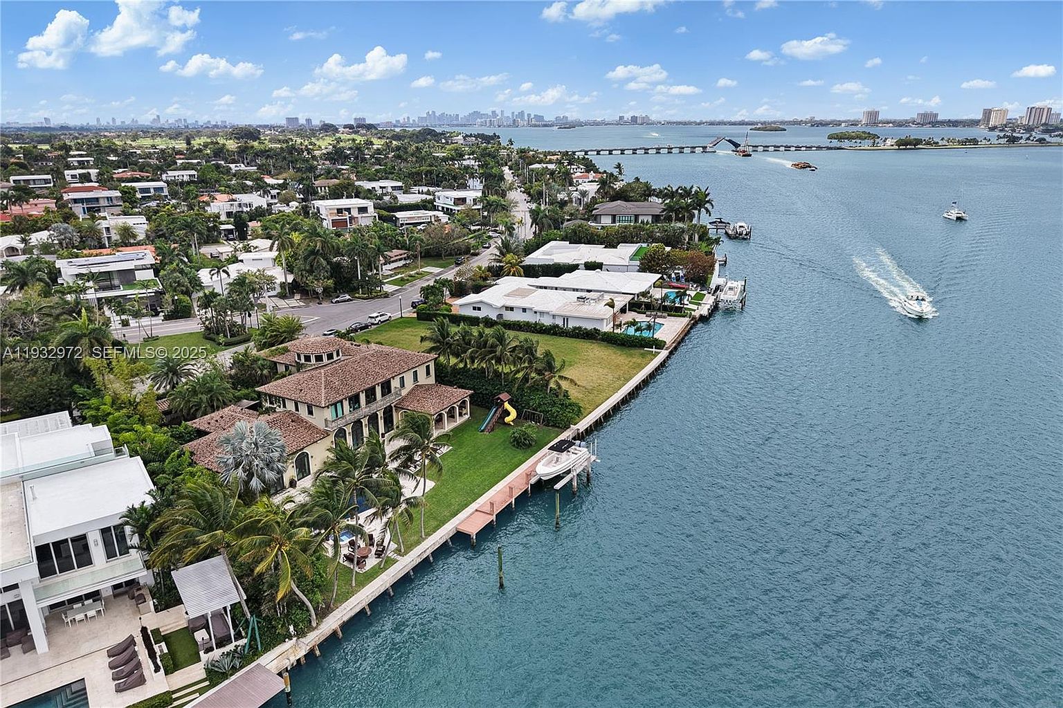 This high-angle aerial view captures a luxurious waterfront property in a coastal residential neighborhood. The scene features a large multi-story home with a tiled roof, a sprawling green lawn, and a private dock extending into the blue waterway, where a boat is actively cruising. In the distance, a bridge spans the water, leading toward a cityscape skyline under a bright, partly cloudy sky.