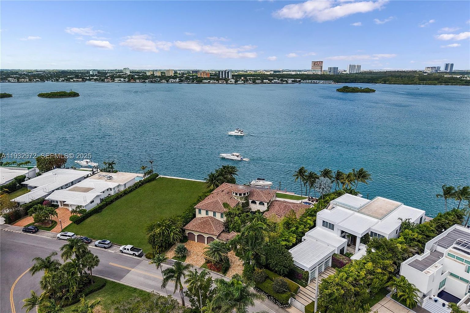 This high-angle aerial view captures a waterfront residential property featuring a Mediterranean-style home with a terracotta tile roof. The expansive lot includes a large, manicured green lawn, mature palm trees, and a private dock extending into the bay. The scene is set against a backdrop of open water, distant city skylines, and a bright blue sky with scattered clouds.