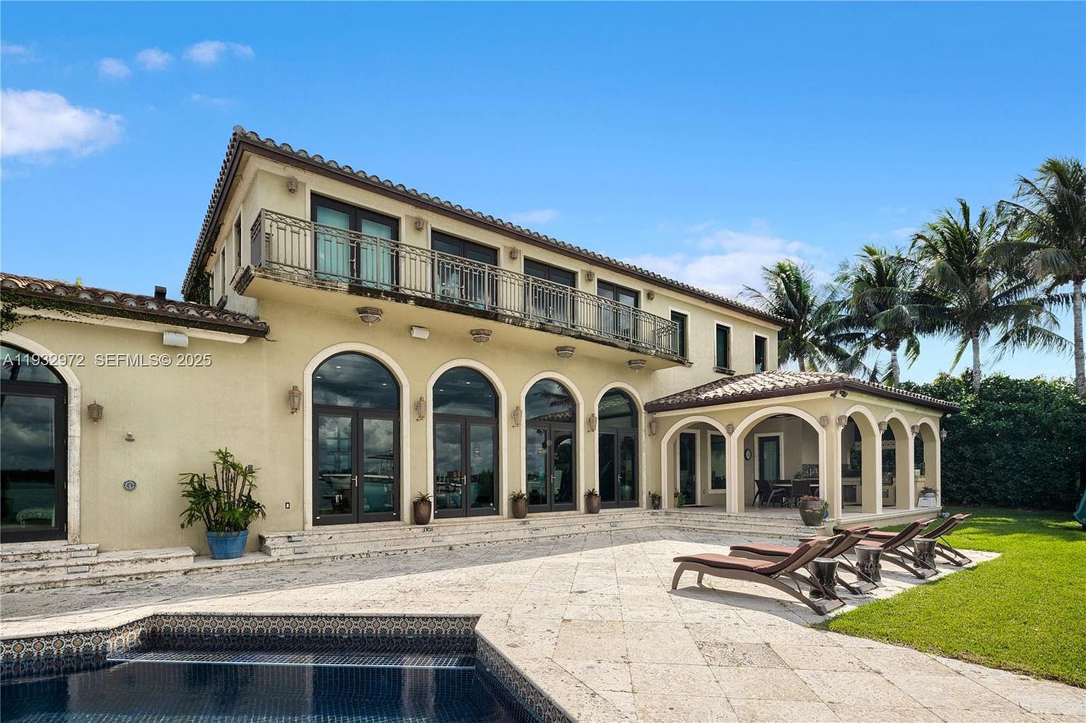This rear view of a Mediterranean-style estate showcases a spacious stone-paved patio area adjacent to a swimming pool. The two-story residence features a series of arched glass doors, a second-story balcony with decorative metal railings, and a covered outdoor living space with arched openings. The scene is set under a clear blue sky, highlighting the property's expansive outdoor entertainment potential.