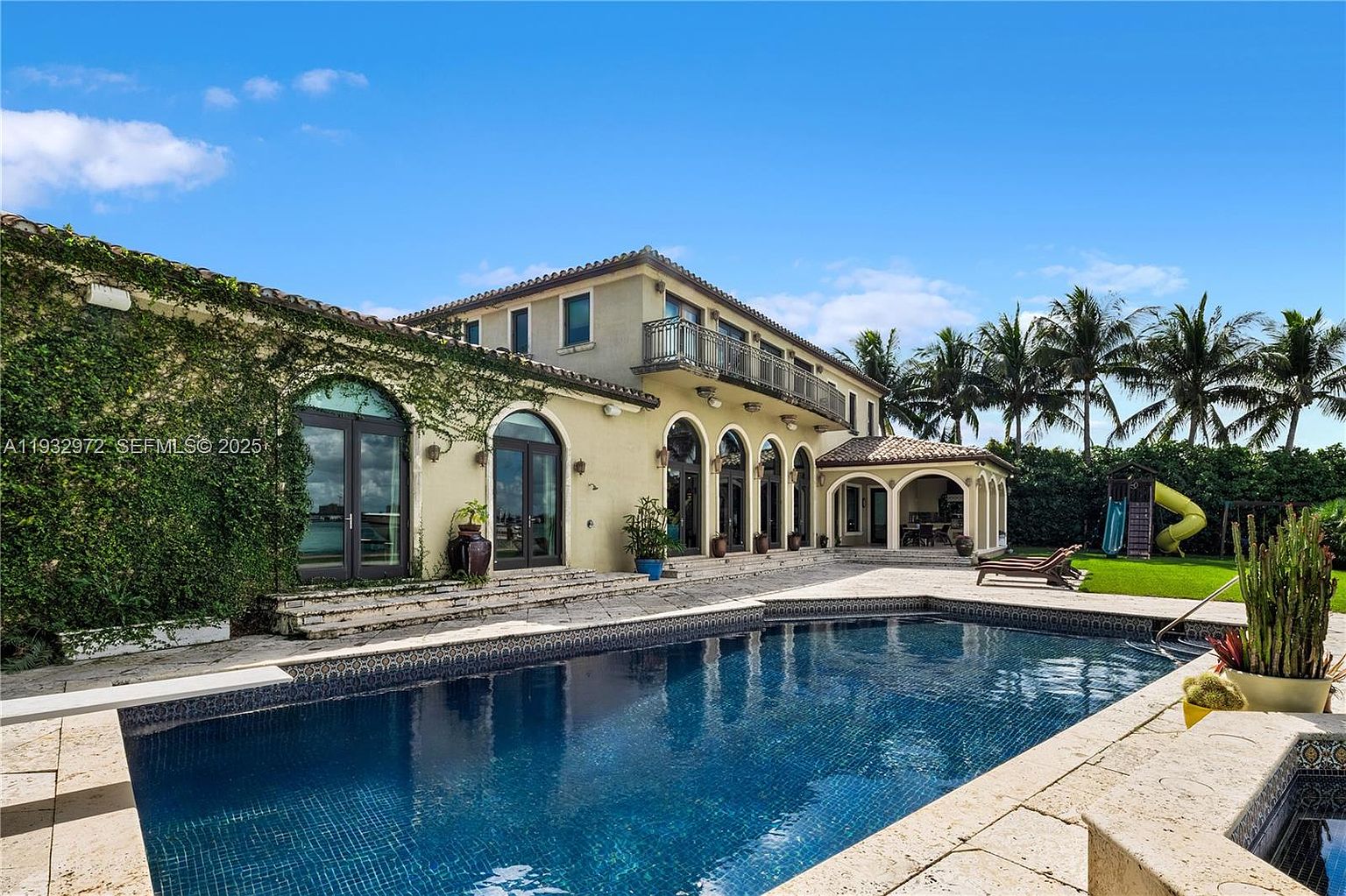 This expansive rear exterior view showcases a Mediterranean-style estate featuring a large, deep blue swimming pool and an adjacent stone-paved patio. The home is characterized by its warm-toned stucco facade, multiple arched glass doors, and a second-story balcony with decorative iron railings. Lush greenery, including climbing vines on the wall and tall palm trees in the background, creates a serene and luxurious resort-like atmosphere.