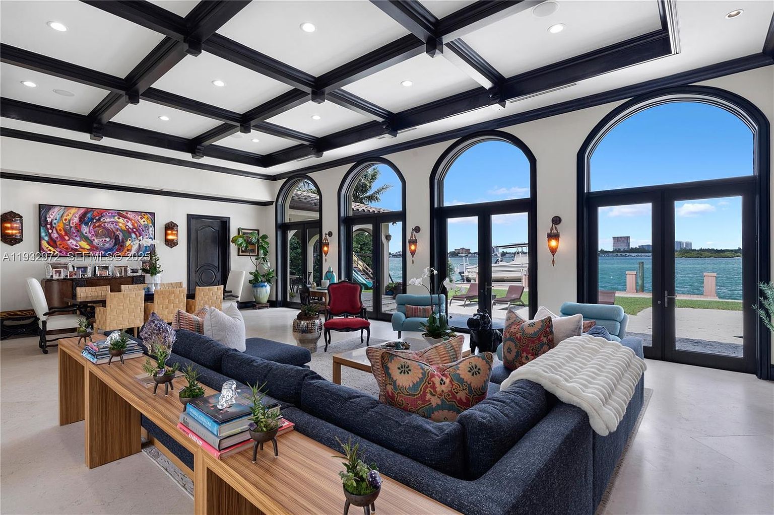 This expansive living room features a coffered ceiling with dark beams and a large, dark-toned sectional sofa arranged to face the water views. Three sets of tall, arched glass doors provide abundant natural light and a seamless connection to the outdoor patio and waterfront. The space is elegantly furnished with a mix of contemporary and classic pieces, including a vibrant abstract painting and a dining area visible in the background.