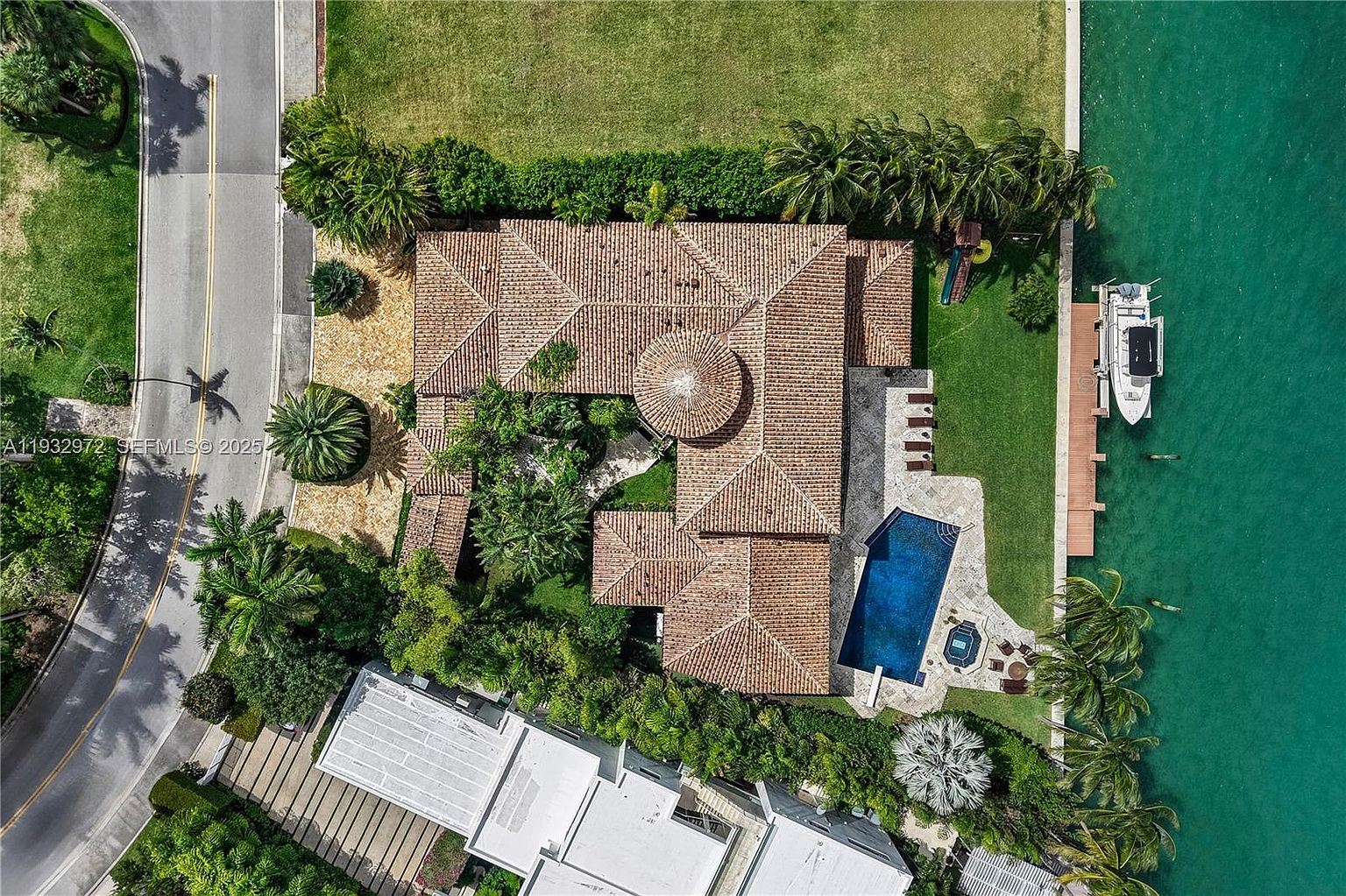 This high-angle aerial perspective captures a sprawling Mediterranean-style residence situated on a waterfront lot. The property features a complex, multi-gabled tile roof, a central circular architectural element, and a private swimming pool with an adjacent spa on the rear patio. A long wooden dock extends into the turquoise water, where a boat is moored, while lush landscaping and mature palm trees frame the manicured lawn.