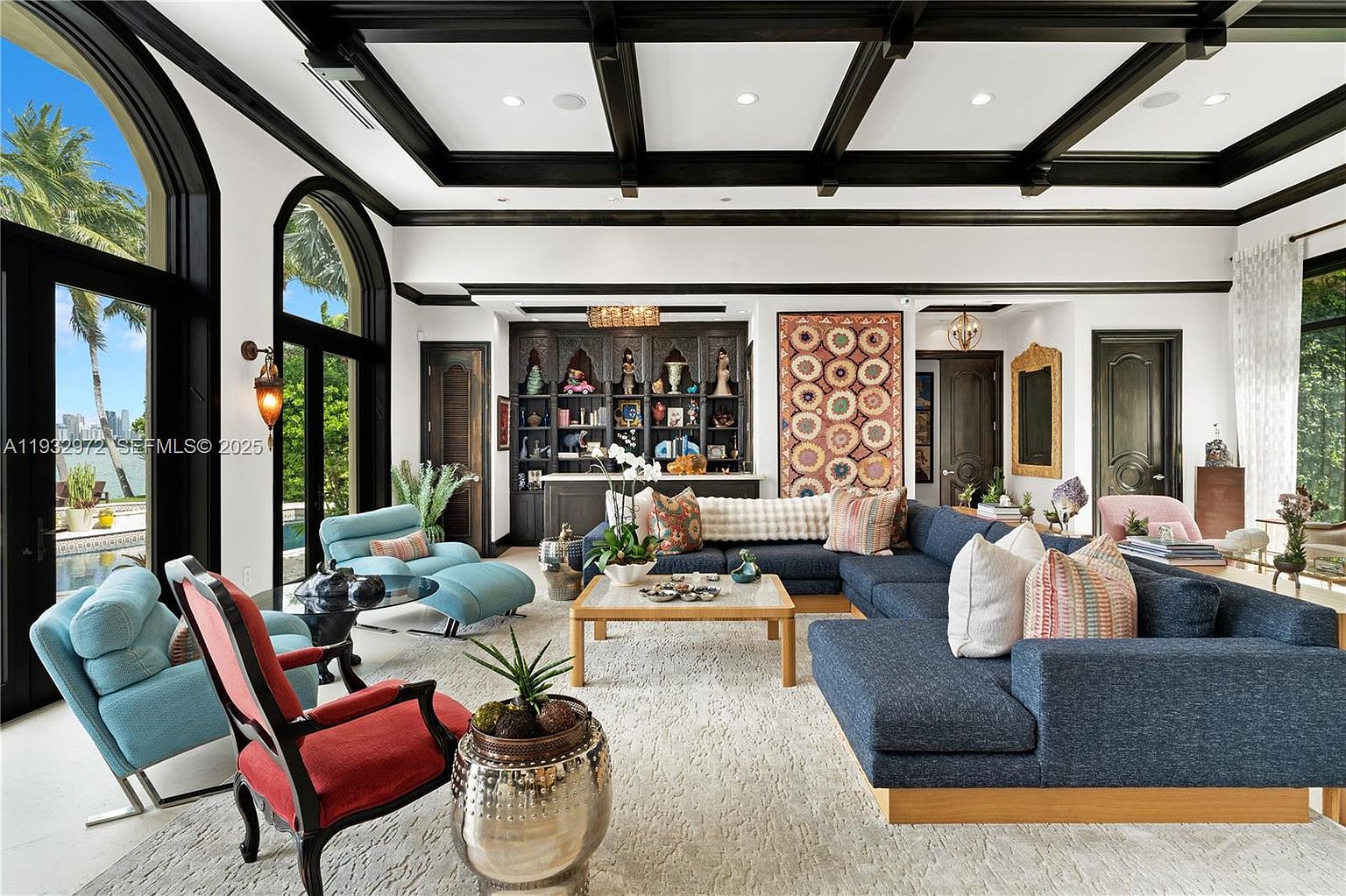 This expansive living room features a coffered ceiling with dark beams and a large, plush blue sectional sofa arranged around a central coffee table. The space is accented by a vibrant, patterned wall tapestry, a built-in bar area with shelving, and a mix of colorful seating including a red-framed chair and light blue armchairs. Large glass doors provide a seamless transition to the outdoor area, filling the room with natural light and offering a view of the pool and distant cityscape.