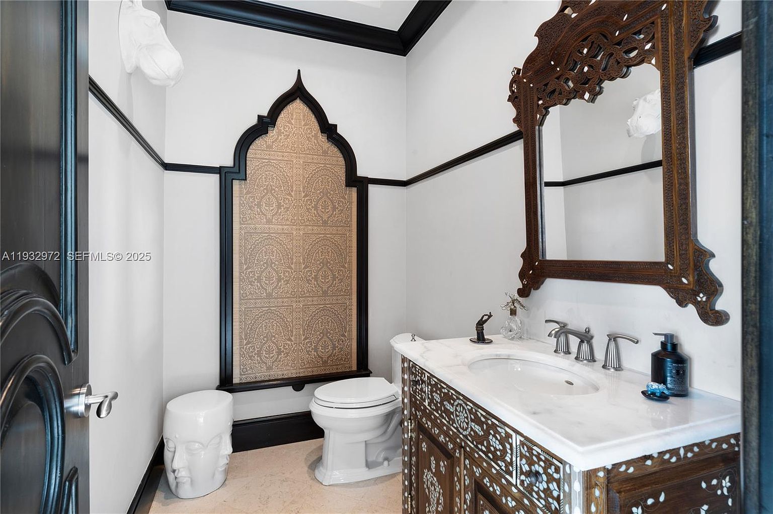 This powder room features a distinct, ornate aesthetic with a dark wood vanity adorned with intricate inlay work and a matching carved mirror frame. A striking arched wall panel with a patterned fabric insert serves as a focal point behind the toilet, complemented by a sculptural white stool. The space is finished with a stone-topped vanity and black trim accents, creating a sophisticated and eclectic atmosphere.