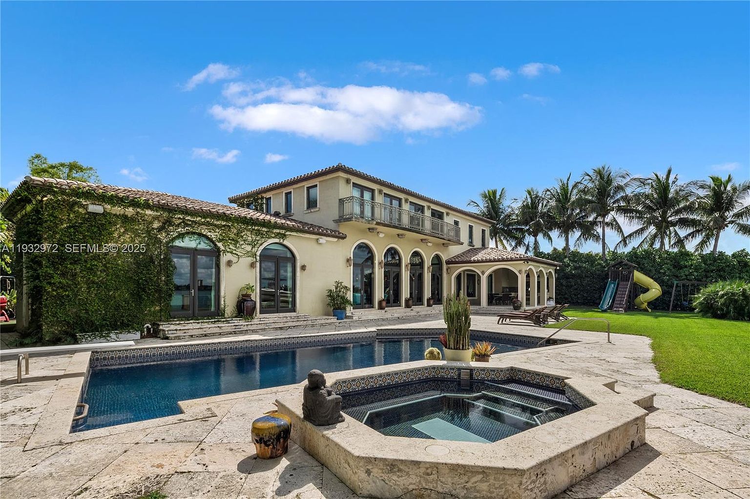 This expansive rear view of a Mediterranean-style estate showcases a large swimming pool and an integrated spa in the foreground. The two-story residence features a light-colored stucco facade, arched glass doors, and a second-floor balcony, all set against a lush green lawn with mature palm trees. The scene captures a serene, resort-like atmosphere perfect for outdoor entertaining.