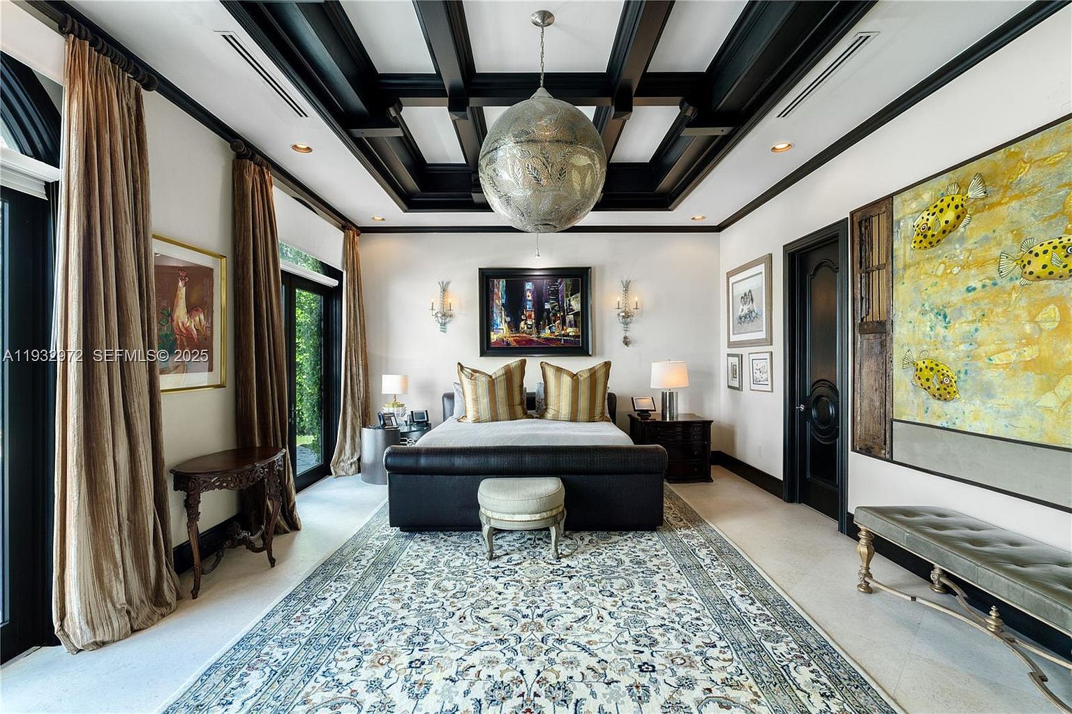 This elegant primary bedroom features a dramatic coffered ceiling with dark trim and a large, ornate metallic pendant light as the centerpiece. The room is anchored by a dark upholstered bed flanked by matching nightstands and wall sconces, all set atop a large, intricately patterned area rug. To the right, a large, vibrant painting of fish adds a bold artistic touch, while tall curtains frame the glass doors leading to the outdoors.