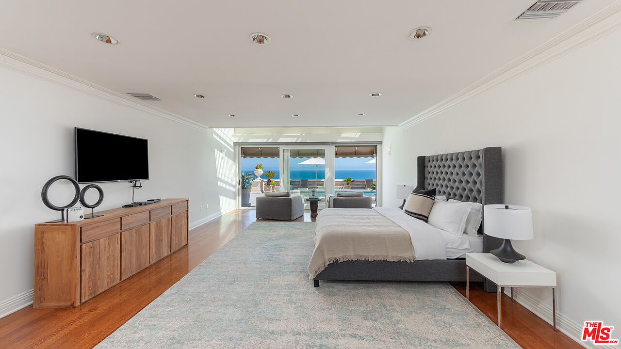 This is a bright and spacious primary bedroom with a view of the ocean. The room features a large bed with a tufted headboard, a wooden dresser with a TV, and a light blue area rug. Sliding glass doors lead to a balcony or patio, enhancing the indoor-outdoor flow.