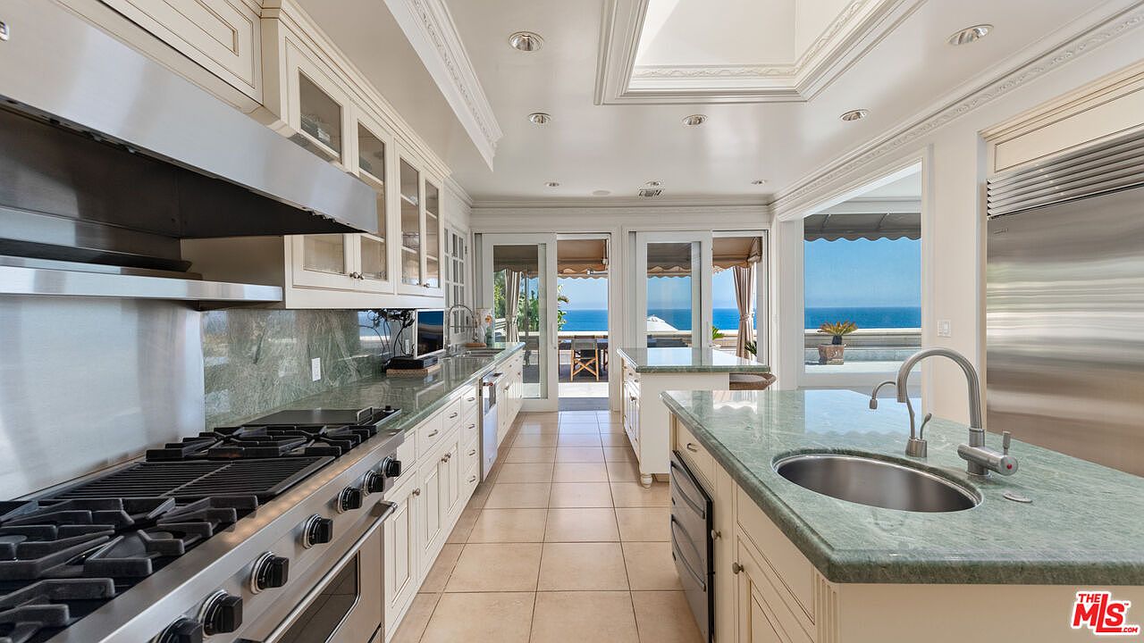 This is a bright and spacious kitchen featuring custom cabinetry, stainless steel appliances, and green marble countertops. The kitchen has a large island with a sink and a view of the ocean through sliding glass doors. The overall style is luxurious and elegant, with a focus on natural light and high-end finishes.