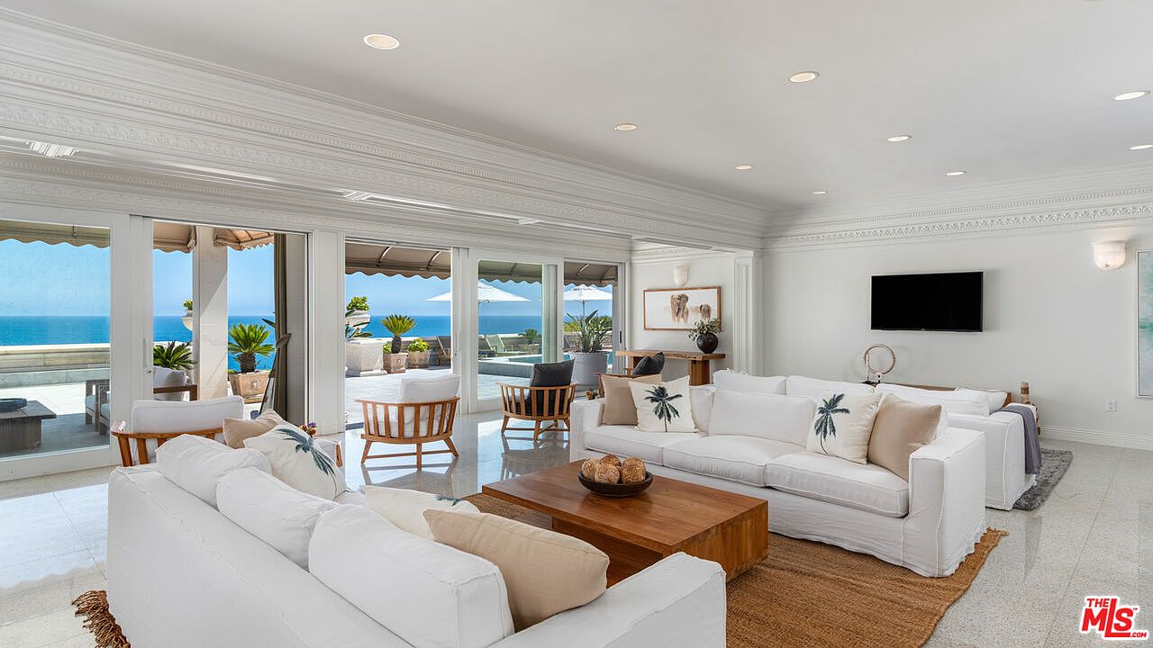 This is an interior shot of a luxurious living room with an ocean view. The room features white sofas, a wooden coffee table, and large sliding glass doors that open to a patio overlooking the ocean. The style is elegant and coastal, creating a bright and airy atmosphere.