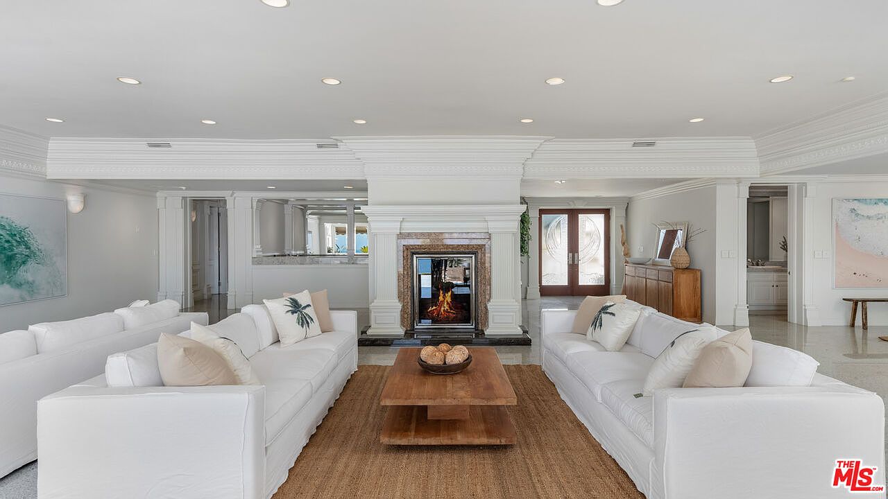 This is an interior shot of a luxurious living room featuring two white sofas facing each other, separated by a wooden coffee table on a jute rug. A fireplace serves as a focal point, and the room is brightly lit with recessed lighting, showcasing the elegant architectural details and creating a welcoming atmosphere. The room also features a bar area and a glimpse into other rooms, suggesting an open floor plan.