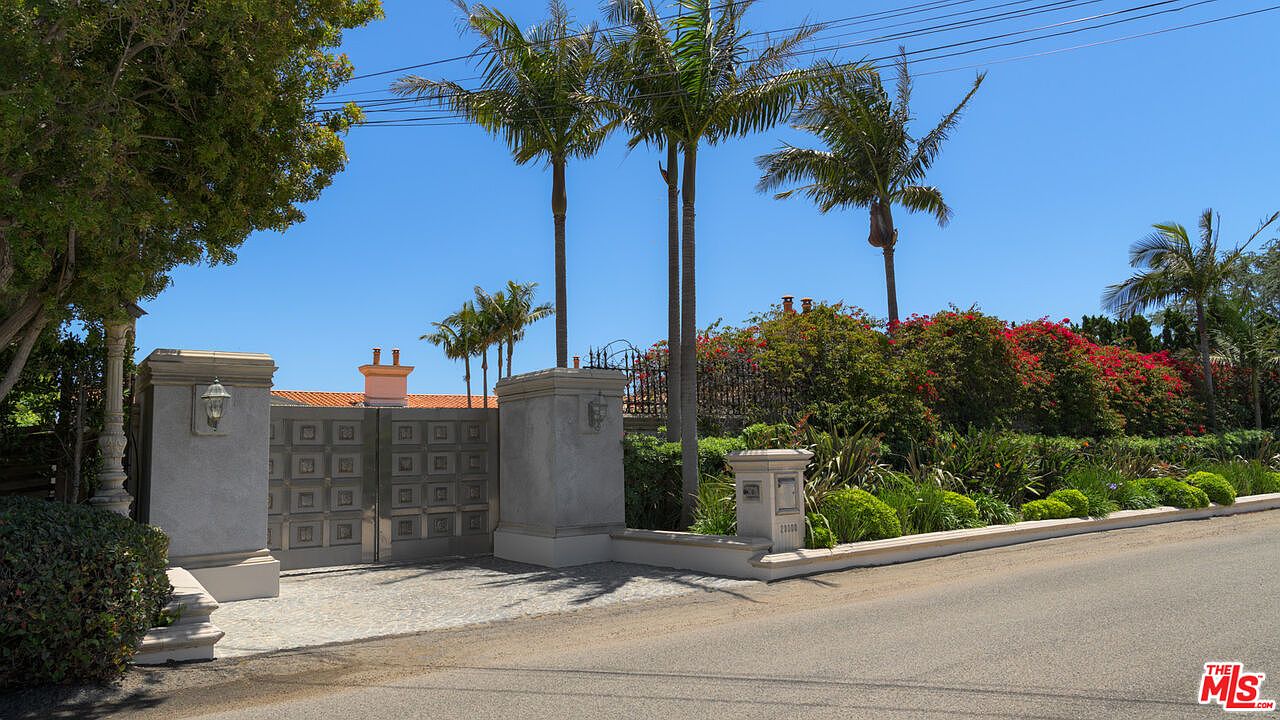 The image showcases an elegant entryway to a property, featuring a gated entrance with substantial stone pillars and decorative gates. Lush landscaping, including palm trees and vibrant flowering bushes, frames the entrance, adding to the property's curb appeal. The scene is captured from a street-level perspective, emphasizing the grandeur and privacy of the estate.