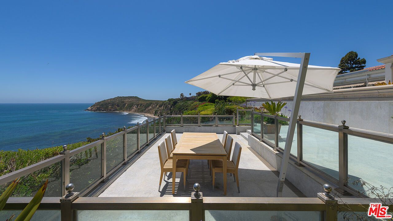 This image showcases a stunning outdoor patio or balcony area with breathtaking ocean views. The space is furnished with a wooden dining table and chairs, sheltered by a large white umbrella. Glass railings provide unobstructed views of the coastline, creating an inviting and luxurious outdoor living space.