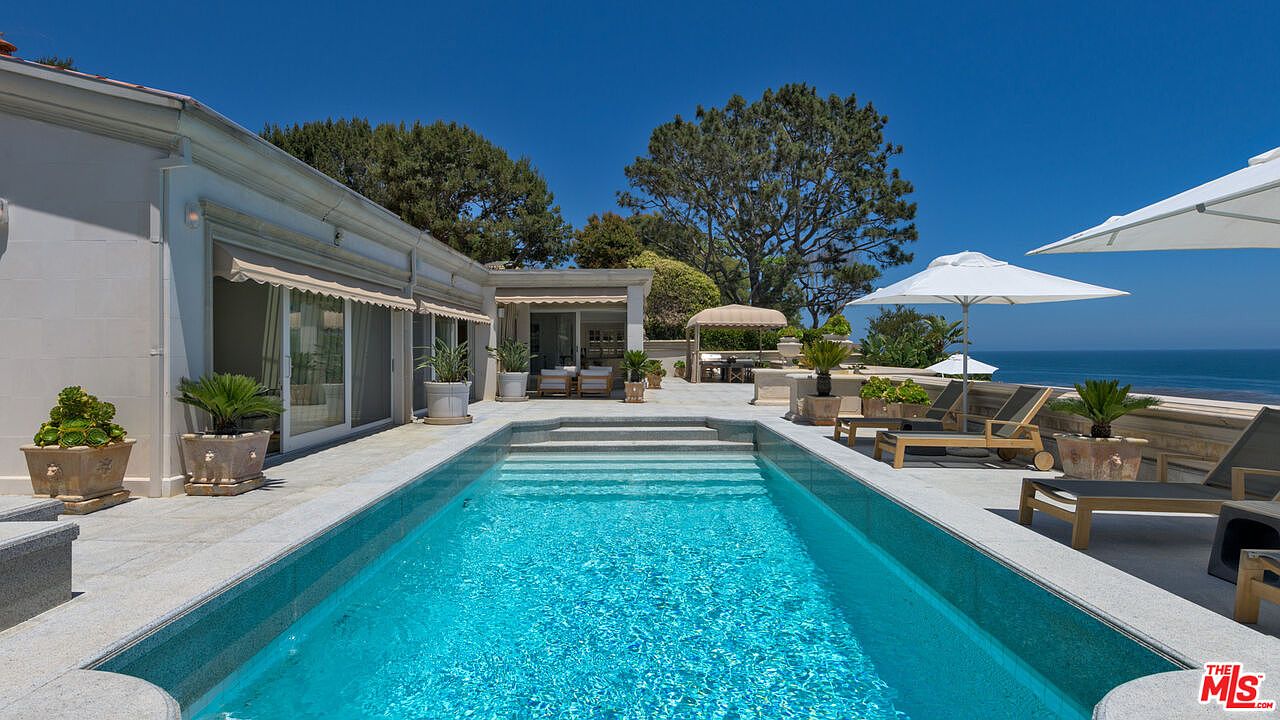 This image showcases a luxurious outdoor pool area with a rectangular pool featuring steps leading into the water. The pool is surrounded by elegant patio furniture, potted plants, and umbrellas, creating a serene and upscale atmosphere. In the background, the ocean is visible, enhancing the property's appeal and suggesting a high-end coastal lifestyle.